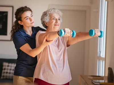 Old woman training with physiotherapist using dumbbells at home. Therapist assisting senior woman with exercises in nursing home. Elderly patient using dumbbells with outstreched arms in a physical therapy session in clinic.