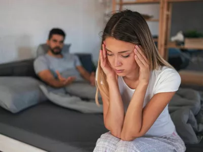 Beautiful depressed woman sitting on bed while her angry boyfriend yells at the background