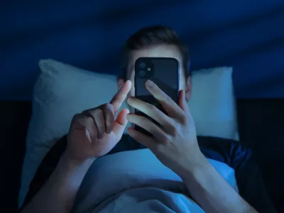 Close up of young man, alone in the dark bedroom, holds his smartphone close to face while scrolling social media or texting late at night. Screen dependence, excessive consumption of content