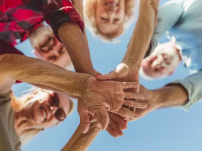 Low angle view of group of cheerful active senior people enjoying sunny summer day outdoors, gathered in circle, holding hands all together in the middle