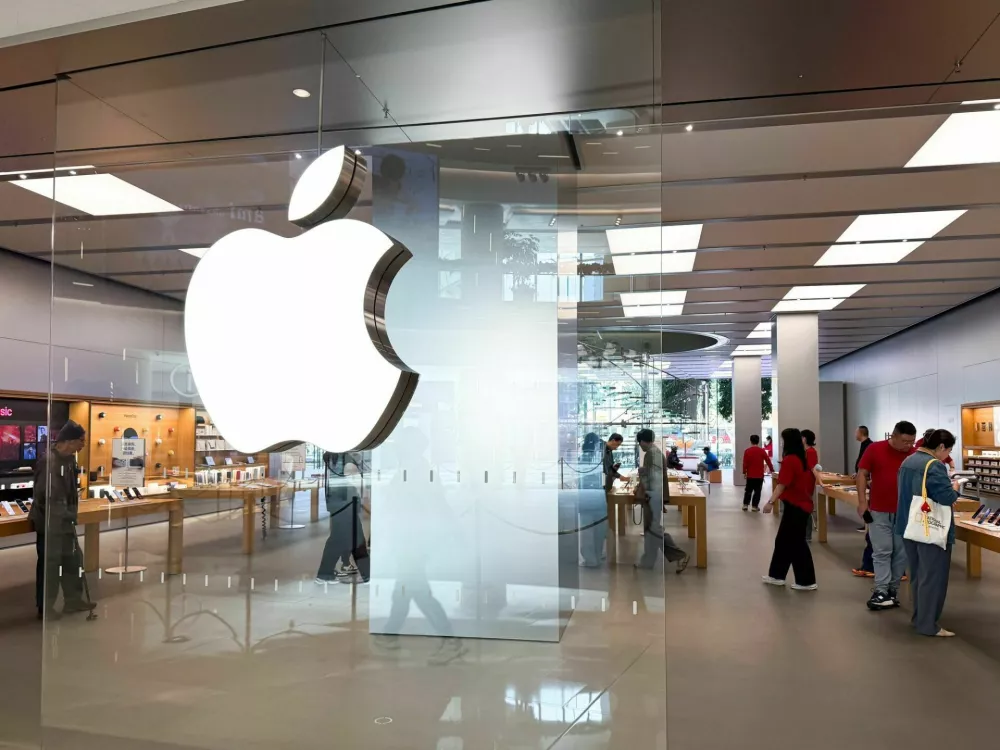 Shanghai, China - November 24, 2025: View through the glass entrance of an Apple Store in Shanghai showing the illuminated Apple logo, customers browsing products, Apple staff in red uniforms, wooden display tables and products in a bright modern retail space. / Foto: Anatoly Kireev