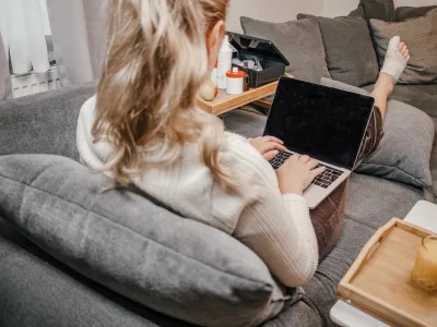 Wide angle shot of a woman resting on a comfortable sofa with her injured, bandaged leg elevated while typing on a laptop computer. On her lap tray stands a baby monitor camera, showing multitasking as a mother working from home during injury or sick leave. Modern remote work, freelancing, motherhood, health recovery, flexible workflow, and telecommuting concept. / Foto: Katharina