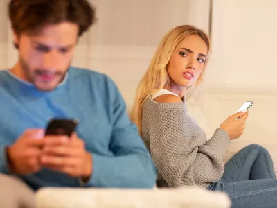 Infidelity. Wife Catching Cheating Husband Texting On Cellphone With Another Woman Sitting On Sofa At Home. Selective Focus