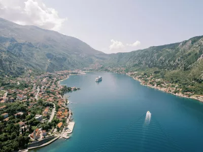 A panoramic aerial photograph of Kotor Bay in Montenegro, showing a cruise ship anchored in the calm blue waters, surrounded by lush green mountains and the historic town of Kotor along the shoreline.