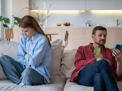 Jealous wife sad about husband messages in phone sitting on couch at home. Spouses ignoring each other. Upset woman turn out from man who looking at smartphone. Betrayal infidelity mistrust concept.