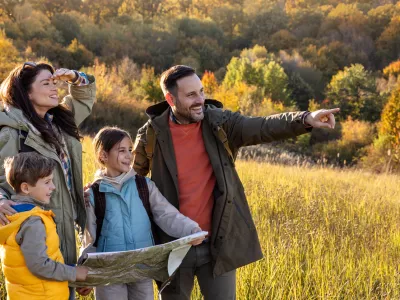 Family standing in sunny field while reading a map, children hold it and father points at viewpoint