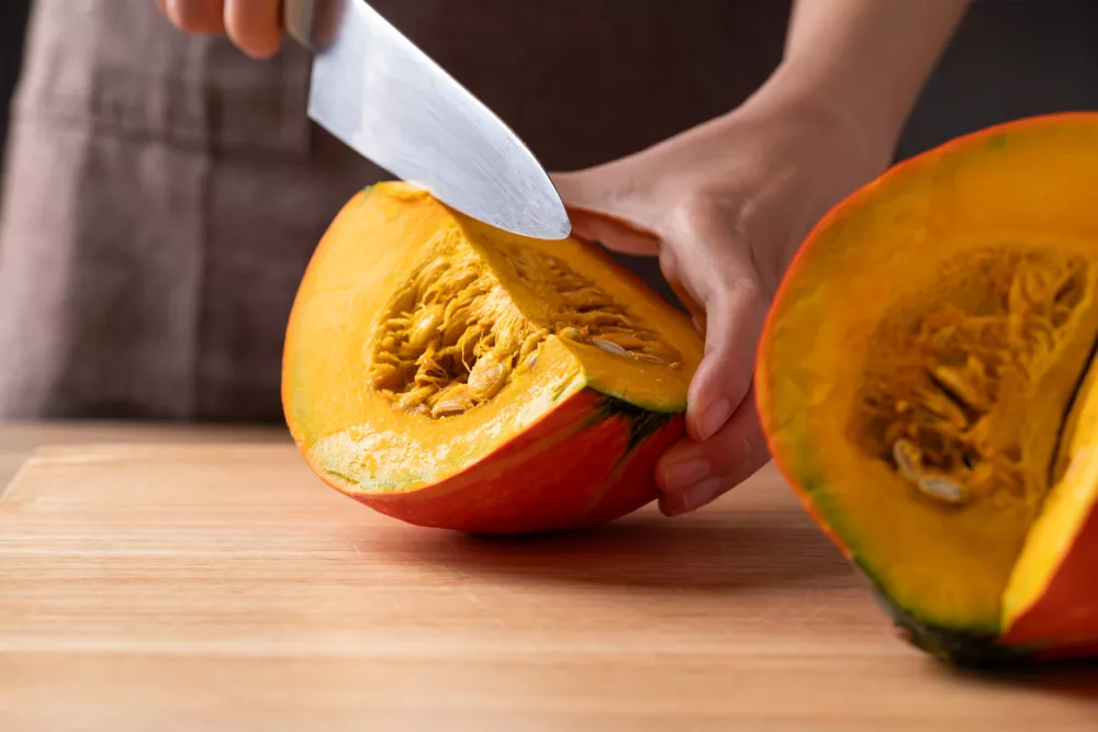 Close-up of hands using a kitchen knife to cut an orange Kuri squash (Hokkaido pumpkin) on a wooden board for homemade cooking