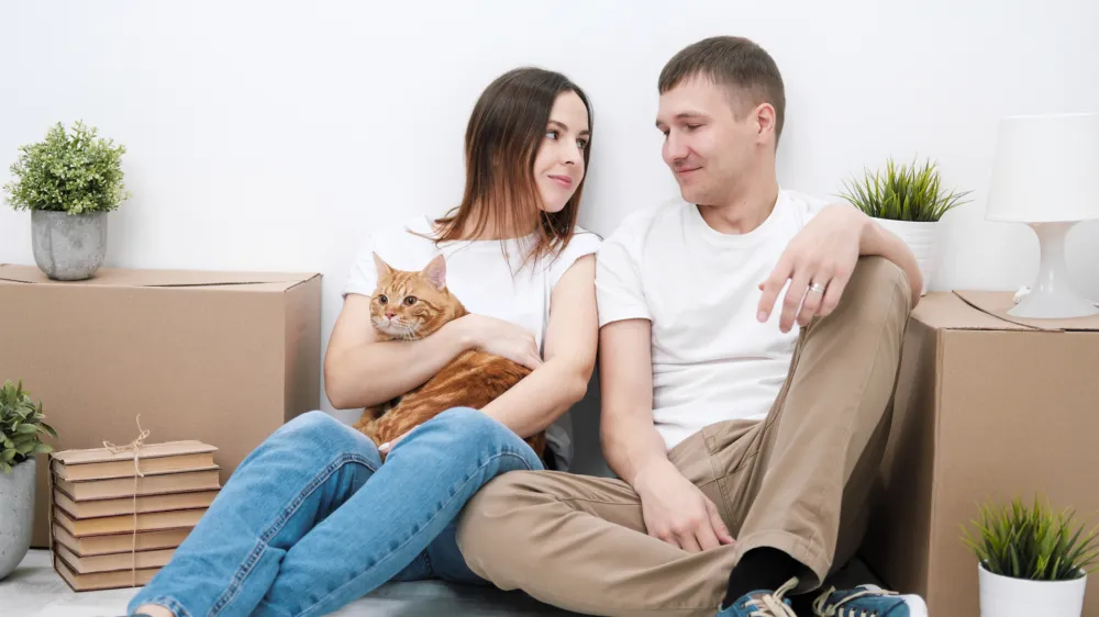 A young married couple with a red cat, a man and a woman, are sitting on the floor in a bright room against the background of cardboard boxes, green plants and things. Concept themes moving to a new home.