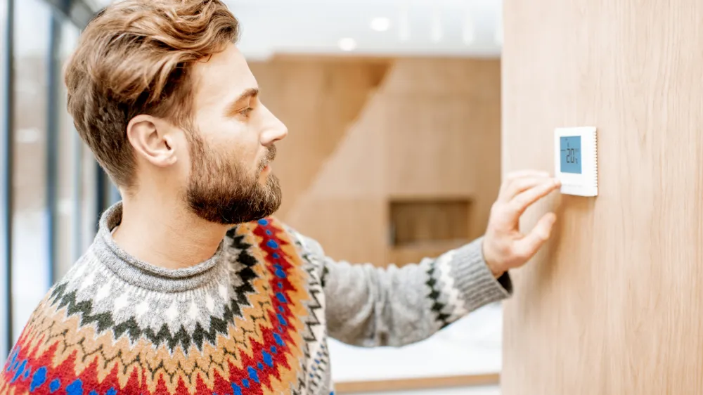 Man in sweater feeling cold adjusting room temperature with electronic thermostat at home