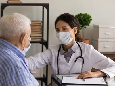 Young female GP doctor in face mask giving support and safe from infection consultation to senior 80s patient, touching shoulder at appointment. Elderly healthcare, medic care, coronavirus concept