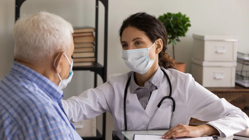 Young female GP doctor in face mask giving support and safe from infection consultation to senior 80s patient, touching shoulder at appointment. Elderly healthcare, medic care, coronavirus concept