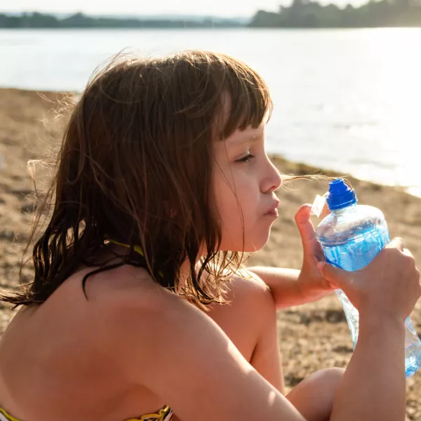 A girl drinks water while sitting on the beach by the lake during a summer day. / Foto: Marizza