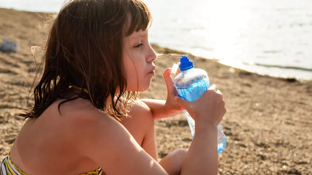 A girl drinks water while sitting on the beach by the lake during a summer day. / Foto: Marizza