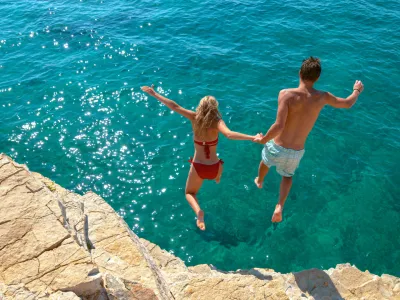 Carefree tourists hold hands while jumping into the refreshing blue sea during a relaxing summer vacation. Active young woman and her boyfriend dive off a high cliff and into the deep blue ocean.