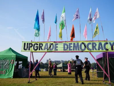 30 August 2024, United Kingdom, Windsor: Police officers walk beneath a banner reading 'upgrade democracy', among tents in Windsor Great Park, as Extinction Rebellion stage a mass occupation. Activists are calling for the Government to create and be led by a citizens' assembly on climate and ecological justice. Photo: Aaron Chown/PA Wire/dpa