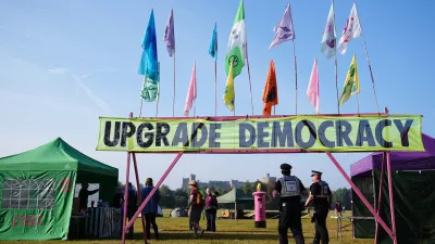 30 August 2024, United Kingdom, Windsor: Police officers walk beneath a banner reading 'upgrade democracy', among tents in Windsor Great Park, as Extinction Rebellion stage a mass occupation. Activists are calling for the Government to create and be led by a citizens' assembly on climate and ecological justice. Photo: Aaron Chown/PA Wire/dpa