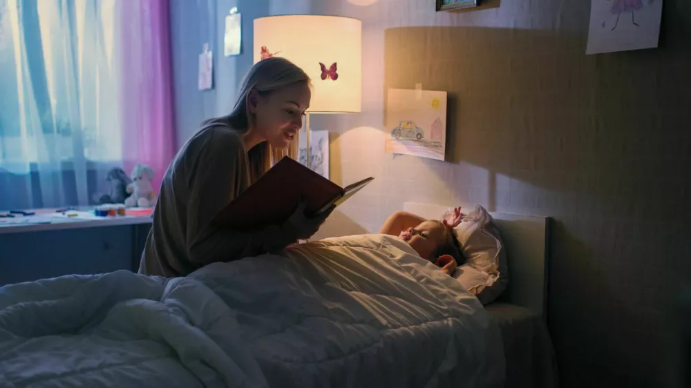 Young Loving Mother Reads Bedtime Stories to Her Little Beautiful Daughter who Goes to Sleep in Her Bed. / Foto: Gorodenkoff