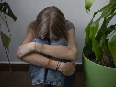 A teenage girl sits with her arms clasped on her knees on the floor against a wall next to houseplants. She's upset and sad. / Foto: Nina Borisova