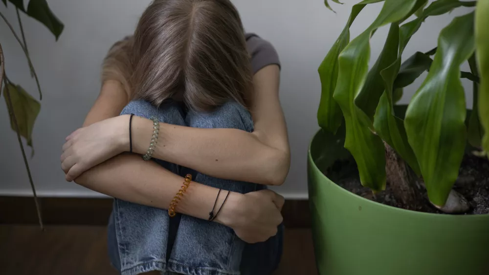 A teenage girl sits with her arms clasped on her knees on the floor against a wall next to houseplants. She's upset and sad. / Foto: Nina Borisova