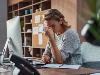 ﻿Exhausted businesswoman having a headache in modern office. Mature creative woman working at office desk with spectacles on head feeling tired. Stressed casual business woman feeling eye pain while overworking on desktop computer.