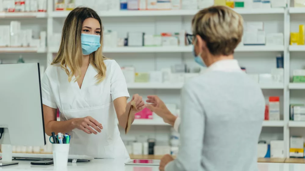 Female pharmacist wearing protective mask and serving a customer patient in a pharmacy and packing drugs in a paper bag