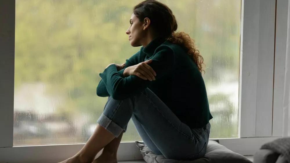 Pensive sad millennial Hispanic woman sit on windowsill at home look in window distance thinking dreaming. Thoughtful young Latino female loner or outcast feel lonely relax rest indoors on rainy day. / Foto: Fizkes