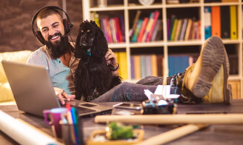 Handsome tattooed man working at home on laptop while sitting at the table with cute dog