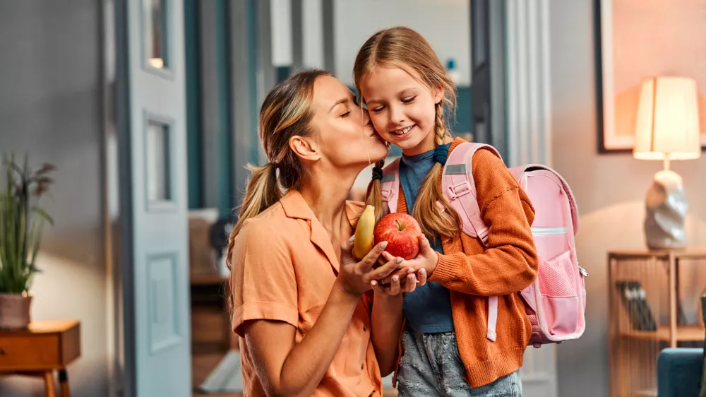 Healthy snack for pupil. Adult woman giving pretty daughter with pink schoolbag apple and banana and kissing her in cheek. Casual female taking care about nutritious meal between lesson for child. / Foto: Harbucks