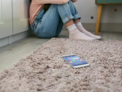 Mobile phone lying on the floor with unrecognizable sad little girl sitting behind. Selective focus on mobile in foreground / Foto: Doble-d