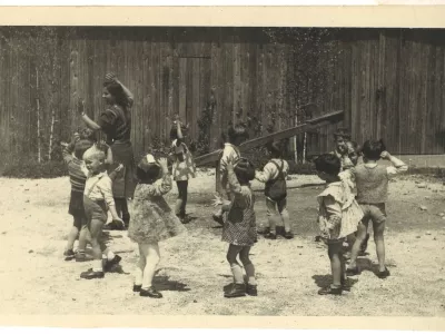 Vrtec Prule, malčki z vzgojiteljico Savico Lanc leta 1956 nastopajo na igrišču pred vrtcem, barako. Plešejo in predstavljajo medvedke. SI_ZAL_LJU/0840, Vzgojno-varstvena organizacija Prule. 