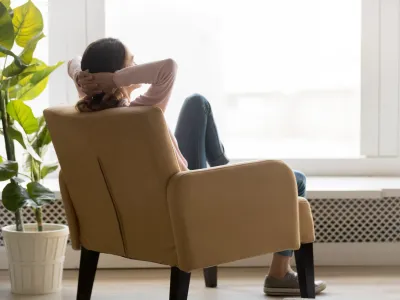 Back view of young happy woman sitting in comfortable armchair, facing shiny window, crossing hands behind head, having rest after housework, resting on weekend, lazy day concept.