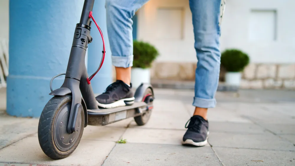 ﻿Close up on woman legs feet standing on the electric kick scooter on the pavement wearing jeans and sneakers in summer day