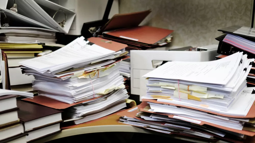 Detail of a messy desk with books binders files and papers stacked up high in a disorganized mess
