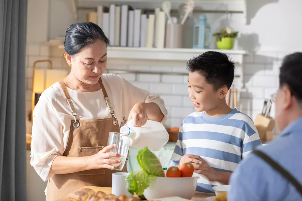Heartwarming scene of Asian grandmother and her grandson in the kitchen. The grandmother is pouring milk into a glass while the grandson watches with smile. Happiness moment