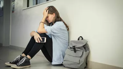 Upset and depressed girl holding smartphone sitting on college campus floor holding head. University sad student suffering from depression sitting on floor at high school. Lonely bullied teen in difficulty with copy space.