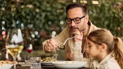 Father and young daughter share a relaxed lunch at an outdoor restaurant, passing food and enjoying quality family time in a casual, warm setting ideal for lifestyle, parenting, and hospitality stories. / Foto: Nortonrsx Getty Images