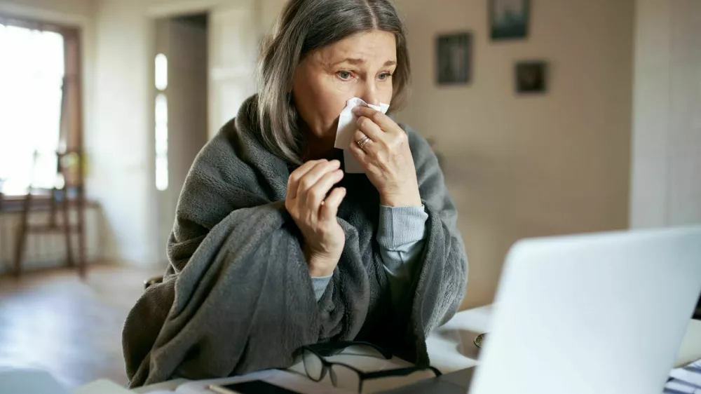 Sick elderly woman wrapped in plaid coughing, sneezing, having respiratory disease symptoms, sitting in front of open laptop, having online video consultation with her physician. Health and technology / Foto: Shurkin_son
