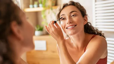 Beautiful woman using mascara on eyelash in bathroom in the morning time. Smiling young woman applying eye make up and looking at mirror. Beauty girl applying black mascara in bathroom at home.