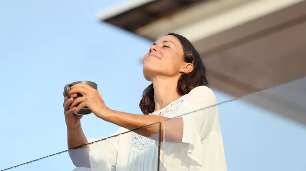 Adult woman with coffee cup breathing on a balcony / Foto: Pheelings Media Getty Images