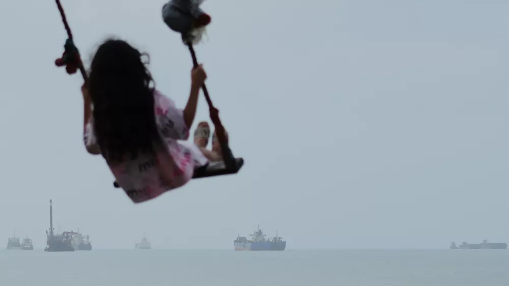 A girl swings along the shore as cargo ships and other commercial vessels appear anchored in the Strait of Hormuz off Bandar Abbas, Iran, Monday, July 27, 2026. (Razieh Poudat/ISNA via AP) / Foto: Razieh Poudat