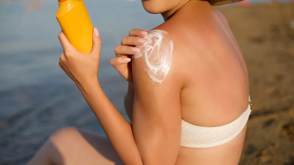 Beautiful woman enjoys a sunny day at the beach while applying sunscreen to her shoulder and preparing for sunbathing, embracing summer skincare rituals near the calm water. / Foto: Verona_s V