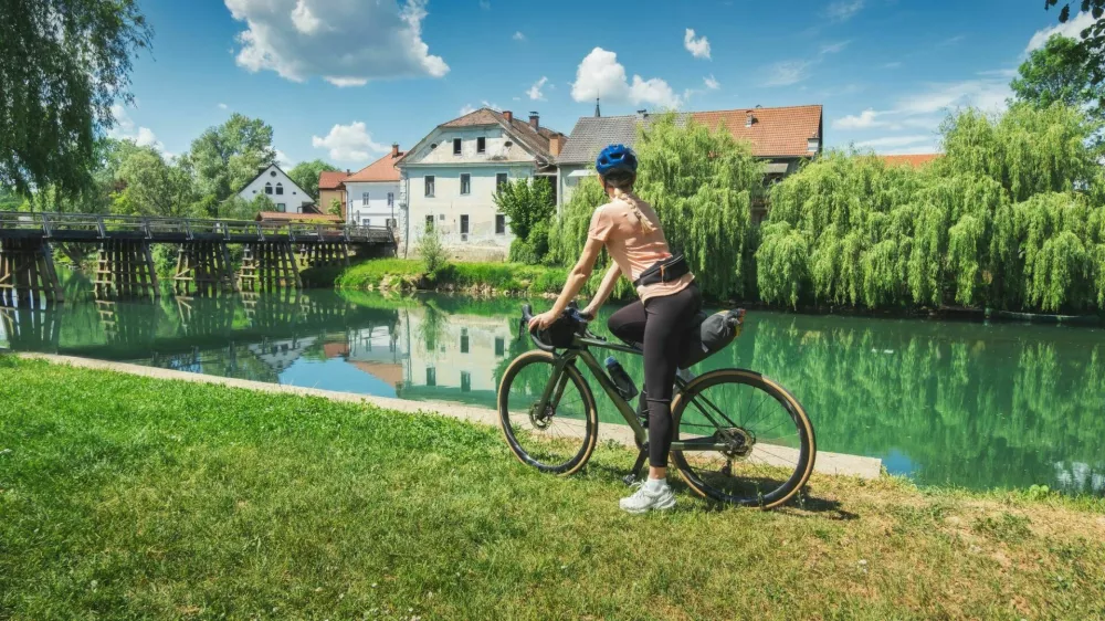 Female cyclist with gravel bike standing on green grass near Krka river in Slovenia. Woman looking at old wooden bridge and traditional village houses. Summer cycling trip, bikepacking. Girl on bike / Foto: Den-belitsky