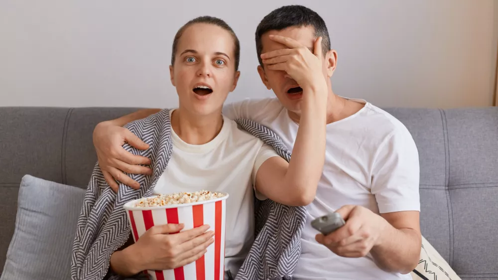 Portrait of impressed woman watching horror film with man, shocked wife covered husband's eyes with palm, forbidden content on tv, couple sitting on sofa wrapped in blanket.