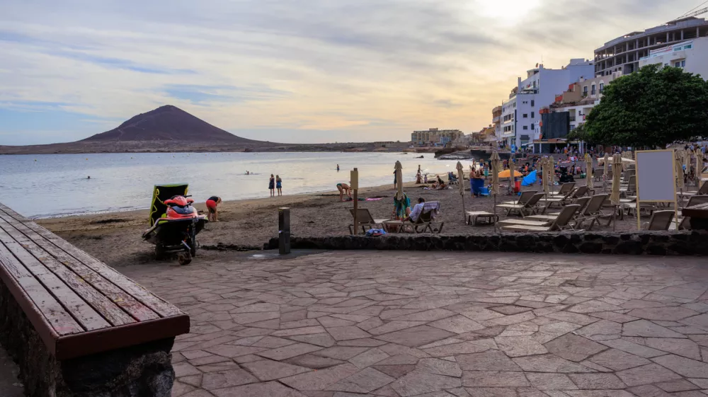 Beach with tourists, sunloungers, sunshades, Atlantic Ocean, hotels and volcanic cone Montana Roja (Red Mountain) at sundown in town El Medano on Canary Island Tenerife, Spain / Foto: Johannes86 Getty Images