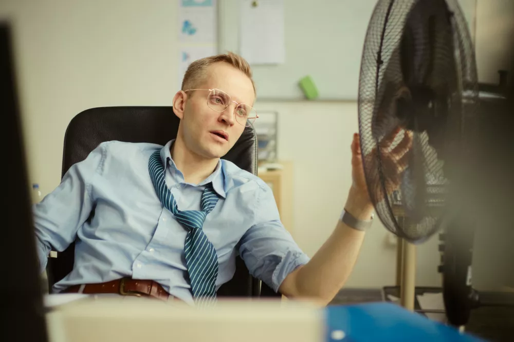 Caucasian young adult businessman cooling himself with electric fan while sitting at desk, showing workplace heat stress and fatigue. Useful for office burnout, summer productivity, employee wellness