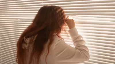 Rear view of beautiful young woman with long hair peeks through hole in the window blinds and looks out the window. Surveillance and curiosity