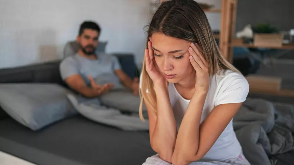 Beautiful depressed woman sitting on bed while her angry boyfriend yells at the background