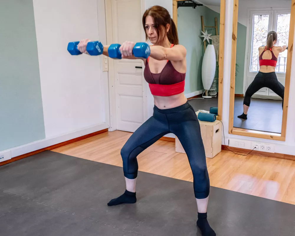 Adult woman performing a front squat exercise with blue dumbbells at home, focusing on strength training and maintaining a healthy lifestyle, with her reflection visible in a mirror / Foto: Alvarort Getty Images