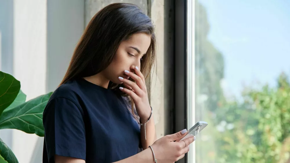 Serious sad teenage female standing at home reading texting on smartphone, copy space. Social life, communication, study education, lifestyle technology concept / Foto: Valeriy_g