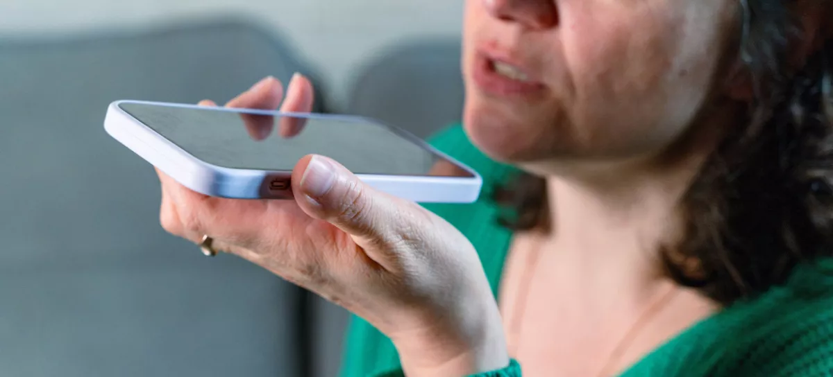 Woman talking on speakerphone with smartphone in hand, seated indoors, wearing a green sweater, with a couch and wall visible in the background / Foto: Andrey Znamenskyi Getty Images